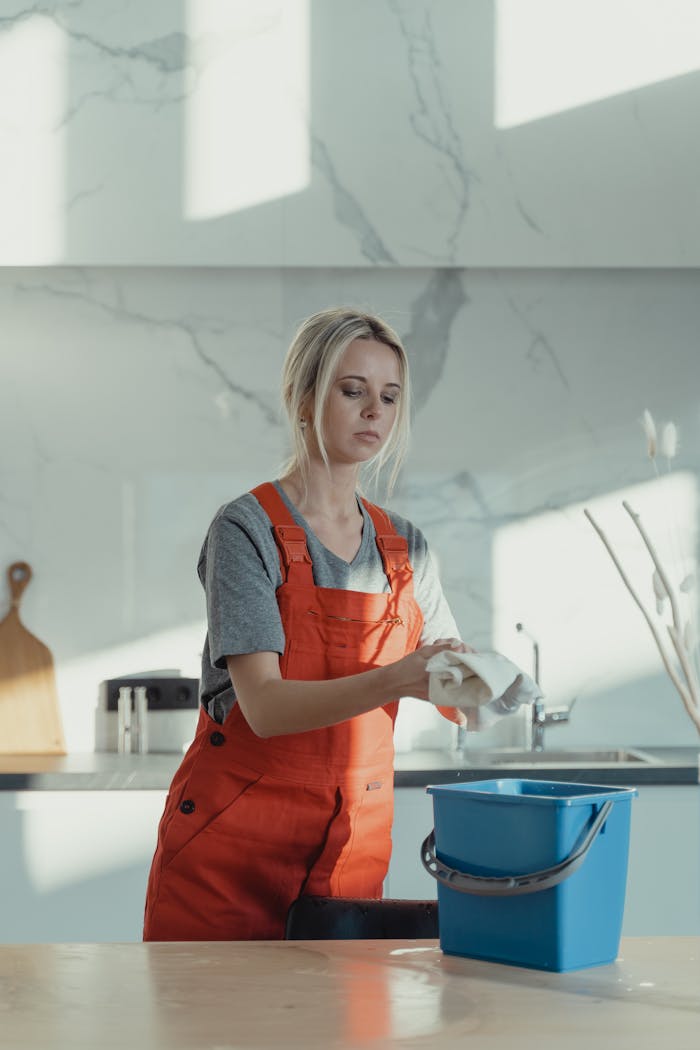 Crafting Captivating Headlines: Your awesome post title goes here A woman wearing orange overalls cleans a kitchen counter with a rag and pail in a modern kitchen.